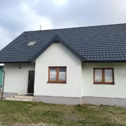 Jednopiętrowy dom z ciemnoszarą dachówką, oknem dachowym i brązowymi ramami okiennymi, świeżo pomalowany na biało.