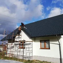 Dwóch pracowników na rusztowaniu przy białym domu z ciemnym dachem, montaż blachy na dachu, widoczne narzędzia i elementy mocujące.