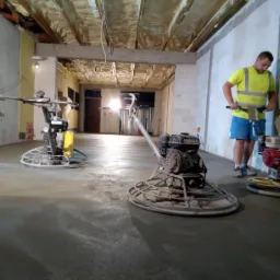 Pracownik w odblaskowej kamizelce wyrównuje świeżo wylaną posadzkę za pomocą zacieraczki mechanicznej, w tle widoczne dwie kolejne maszyny i surowy strop z instalacjami.