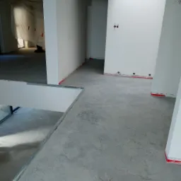 Wnętrze w trakcie wykańczania z szarą, betonową posadzką, zabezpieczone ściany i widoczne instalacje elektryczne.