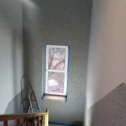 Klatka schodowa w trakcie prac wykończeniowych, ściany częściowo pokryte szarą strukturą, okno zabezpieczone taśmą malarską, drabina, wiadro z farbą i folia ochronna na podłodze.