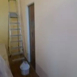 Świeżo pomalowana ściana w jasnym odcieniu, widoczna drabina aluminiowa, wiadro z farbą na podłodze zabezpieczonej folią malarską, drzwi w tle.