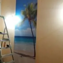 Fragment ściany z naklejoną fototapetą przedstawiającą plażę z palmą, widoczna drabina malarska i narzędzia.