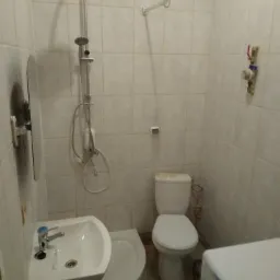 Standardowa łazienka z białą armaturą: umywalka z lustrem, toaleta, kabina prysznicowa z deszczownicą i pralka w rogu, wykończona jasnymi, kwadratowymi płytkami.