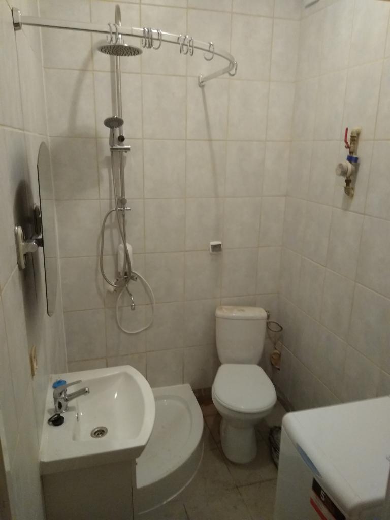 Standardowa łazienka z białą armaturą: umywalka z lustrem, toaleta, kabina prysznicowa z deszczownicą i pralka w rogu, wykończona jasnymi, kwadratowymi płytkami.