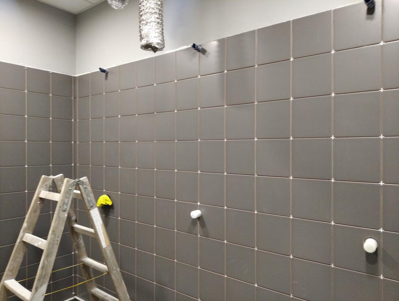 Ściana wyłożona szarymi, kwadratowymi płytkami z białymi krzyżykami dystansowymi, widoczna drewniana drabina i fragment wentylacji podsufitowej.