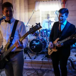 Zespół muzyczny grający na żywo podczas wydarzenia w drewnianym wnętrzu, wokalista z gitarą basową i gitarzysta śpiewają do mikrofonów, w tle perkusista i instrumenty klawiszowe.