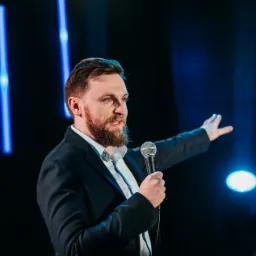 Prelegent z mikrofonem na tle niebieskiego oświetlenia scenicznego w Krakowie, wskazujący ręką w prawo.