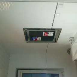 Biała łazienka z panelem rewizyjnym na suficie, widoczna umywalka z baterią i okno.