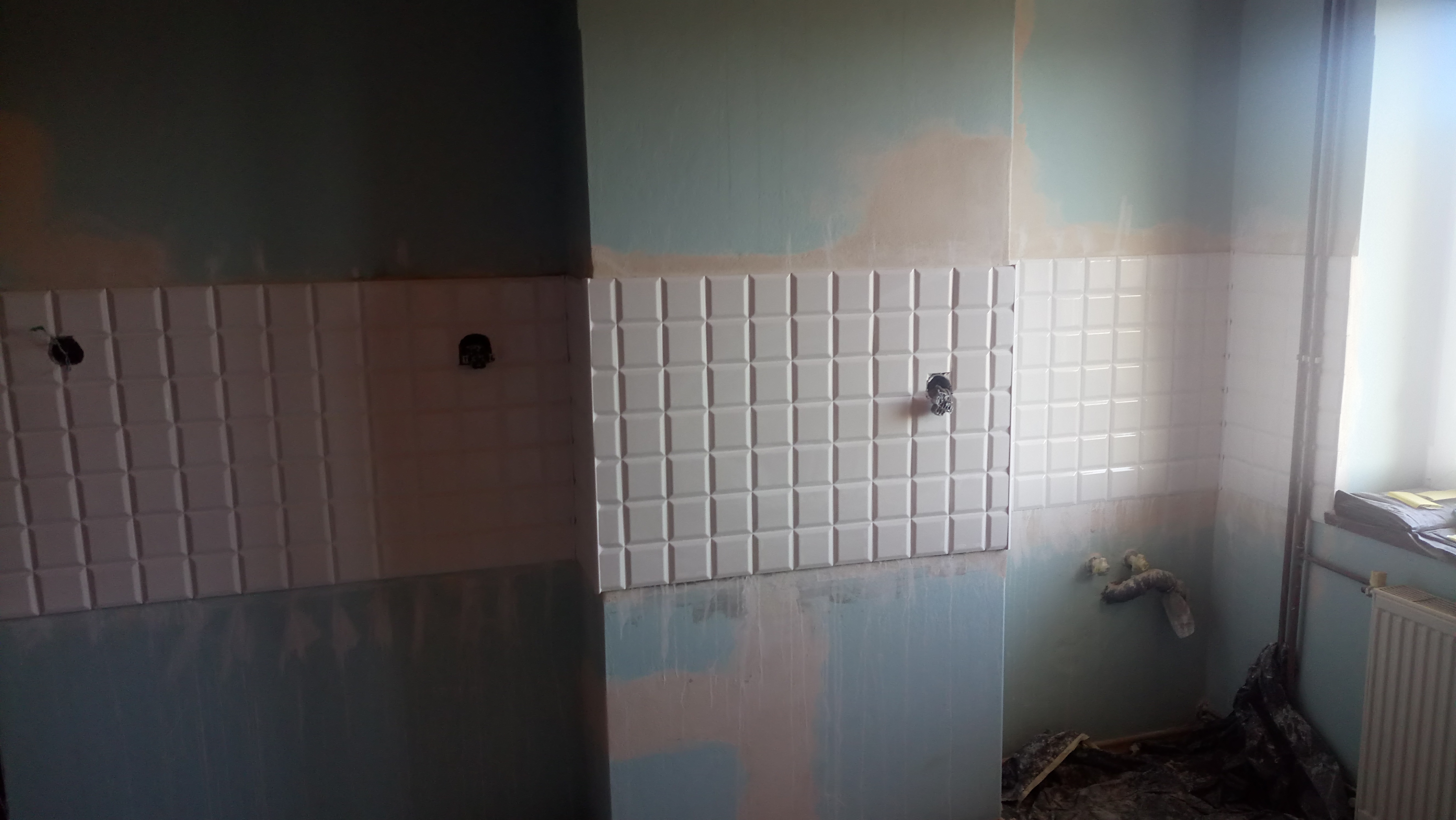 Fragment ściany z częściowo położonymi białymi płytkami kwadratowymi z fugą, widoczne wystające kable i rury, ściana częściowo pomalowana na niebiesko, widoczne ślady po pracach remontowych.