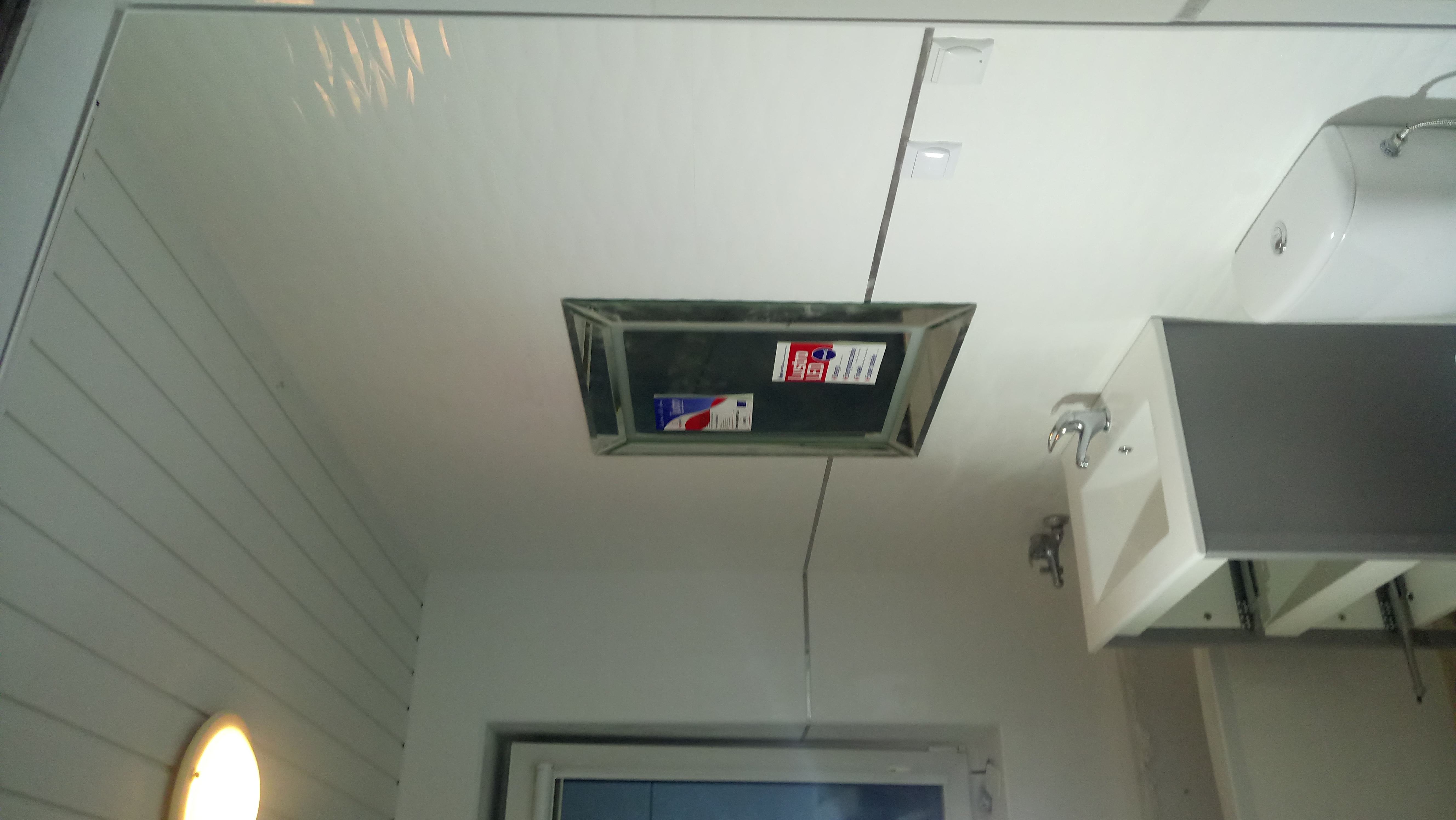 Biała łazienka z panelem rewizyjnym na suficie, widoczna umywalka z baterią i okno.