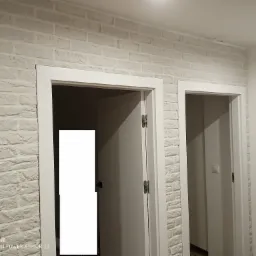 Ściana wykończona dekoracyjną cegłą w odcieniach bieli, obramowująca białe ościeżnice dwóch otwartych drzwi wewnętrznych. Widoczny fragment sufitu z oświetleniem.