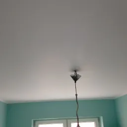 Gładka, biała powierzchnia sufitu z zamontowaną, wiszącą lampą i turkusowymi ścianami z białymi listwami przypodłogowymi.