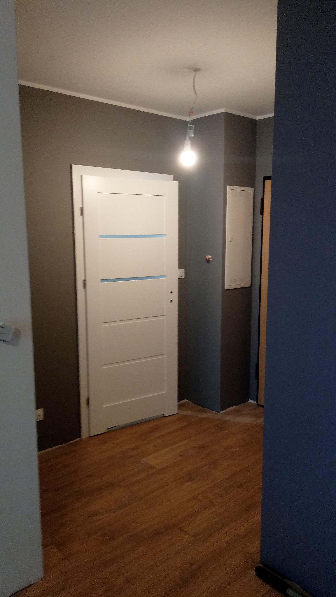 Wnętrze pokoju po remoncie: białe drzwi z poziomymi, niebieskimi paskami na tle szarej ściany, drewnopodobne panele na podłodze, widoczna żarówka bez klosza.
