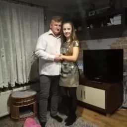 Para na tle ściany z dekoracyjną cegłą w salonie w Żmijewku. Widoczny telewizor i meble. Oświetlenie ciepłe, domowe.