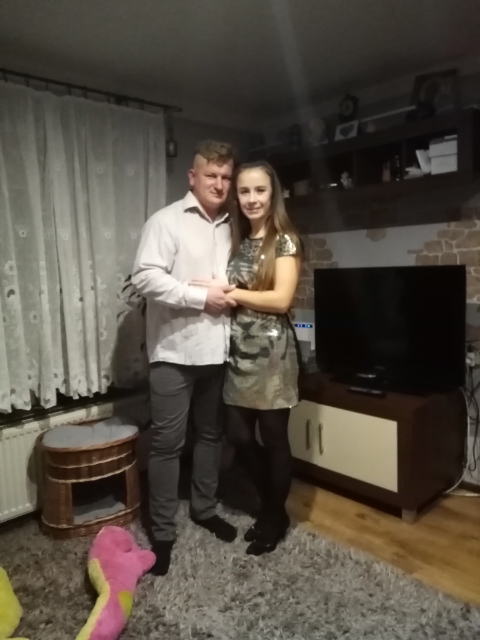 Para na tle ściany z dekoracyjną cegłą w salonie w Żmijewku. Widoczny telewizor i meble. Oświetlenie ciepłe, domowe.