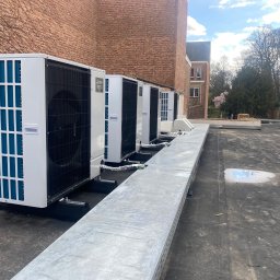 Solar Kontakt - Rząd białych jednostek zewnętrznych klimatyzacji na dachu z widokiem na budynek z cegły i drzewa w tle, instalacja na czarnym dachu pokrytym papą.