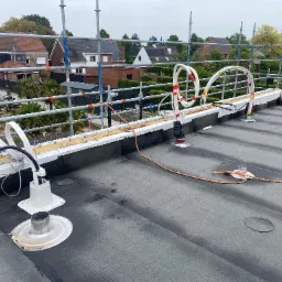 Instalacja na dachu budynku w trakcie montażu, widoczne elementy instalacji, kable i rury. W tle rusztowanie i zabudowa mieszkalna.