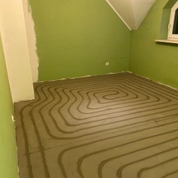 Solar Kontakt - Ułożone rury ogrzewania podłogowego na wylewce w pokoju z zielonymi ścianami i oknem. Widok z góry na spiralny układ rur przed dalszymi pracami wykończeniowymi.