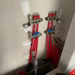 Rozdzielacz ogrzewania podłogowego z czerwonymi rurami, manometrem i zaworami, zamontowany na ścianie w surowym stanie. Widoczne napisy 'WW4' i 'MP' na ścianie.