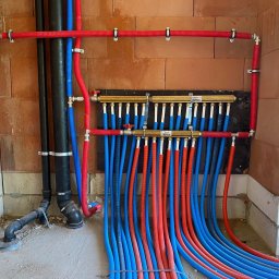 Solar Kontakt - Rozdzielacz ogrzewania podłogowego z rurami w kolorach niebieskim i czerwonym, zamocowany na ścianie z cegieł. Widoczne elementy instalacji w surowym wnętrzu.