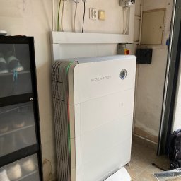 Solar Kontakt - Nowoczesny, biały magazyn energii SIGENergy z pionowymi wentylacjami i subtelnym zielono-czerwonym podświetleniem, podłączony do instalacji elektrycznej na ścianie w pomieszczeniu gospodarczym.