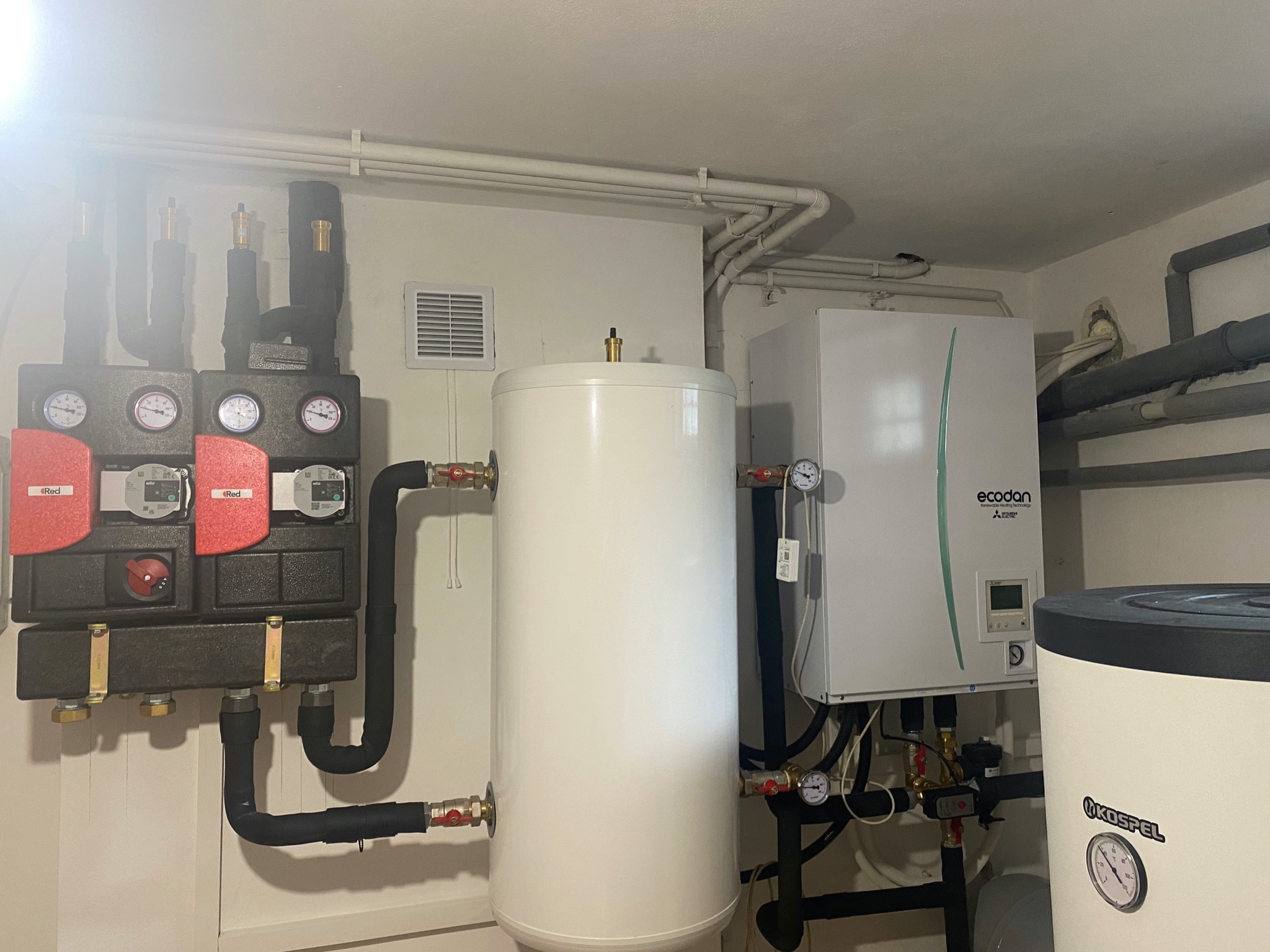 Kompletna instalacja grzewcza w kotłowni: biały zbiornik akumulacyjny Kospel, pompa ciepła Mitsubishi Ecodan z wyświetlaczem, moduł hydrauliczny z czerwonymi pompami obiegowymi Wilo, rury grzewcze...