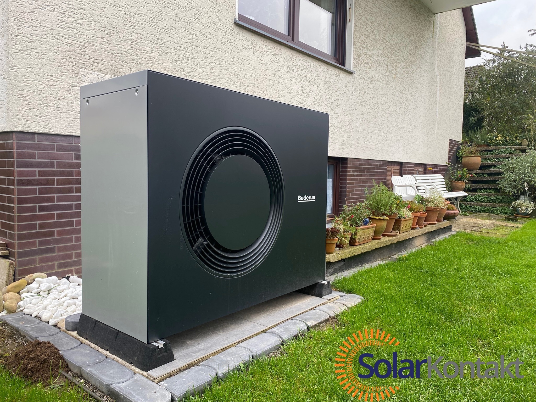Czarna pompa ciepła Buderus, zamontowana przy ścianie domu z beżowym tynkiem, widoczna na tle zielonego trawnika i dekoracyjnych kamieni.