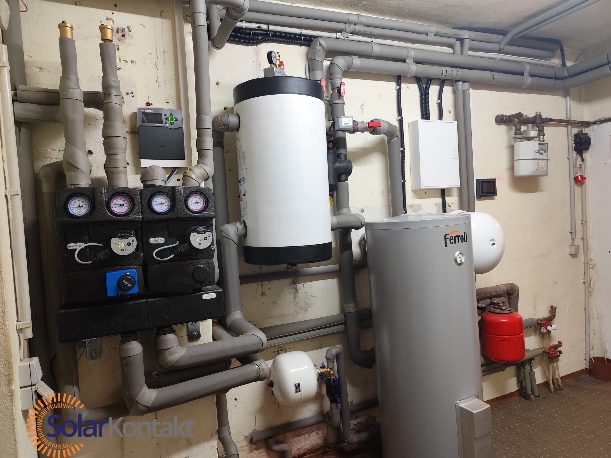 Złożona instalacja grzewcza w piwnicy: zbiornik akumulacyjny Ferroli, liczne rury, zawory i regulatory. Widoczny manometr i logo SolarKontakt w rogu.