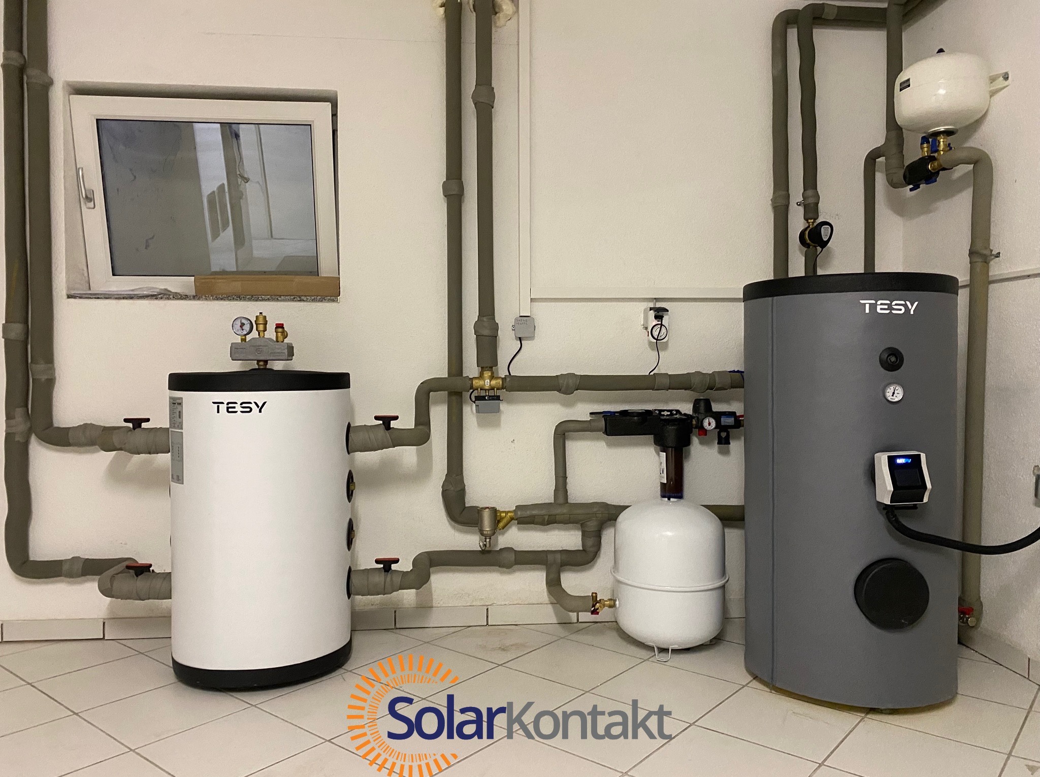 Kompletna instalacja z dwoma zbiornikami Tesy, rurami w izolacji i zaworami, zamontowana w pomieszczeniu z oknem i białymi kafelkami na podłodze.