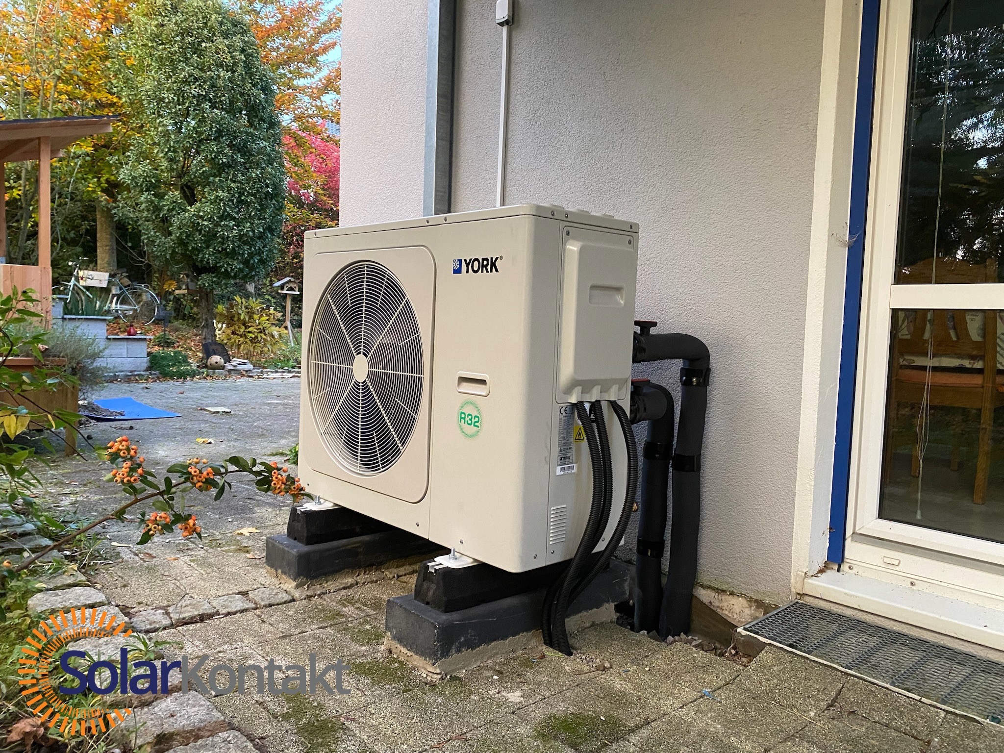 Zamontowana pompa ciepła York na betonowych podstawach, widoczne czarne rury przy elewacji budynku. Tło: jesienne drzewa i fragment ogrodu.
