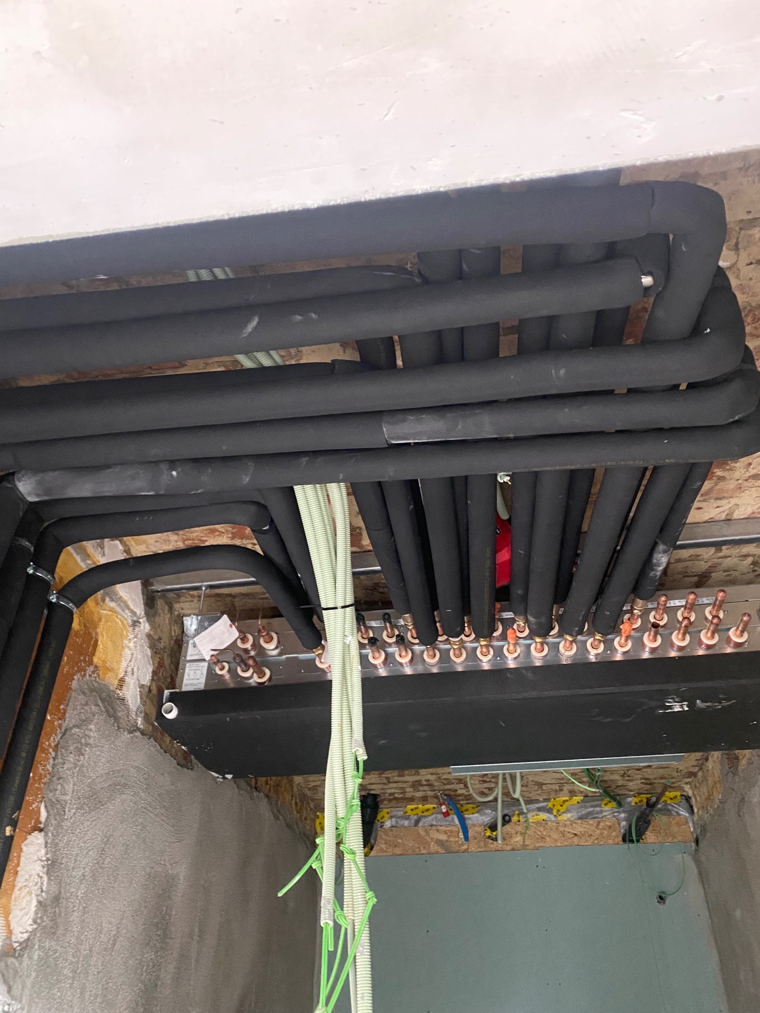 Rozdzielacz ogrzewania podłogowego z rurami w izolacji, widoczne elementy montażowe i instalacja w surowym stanie. Kątowe ujęcie podkreśla złożoność systemu.