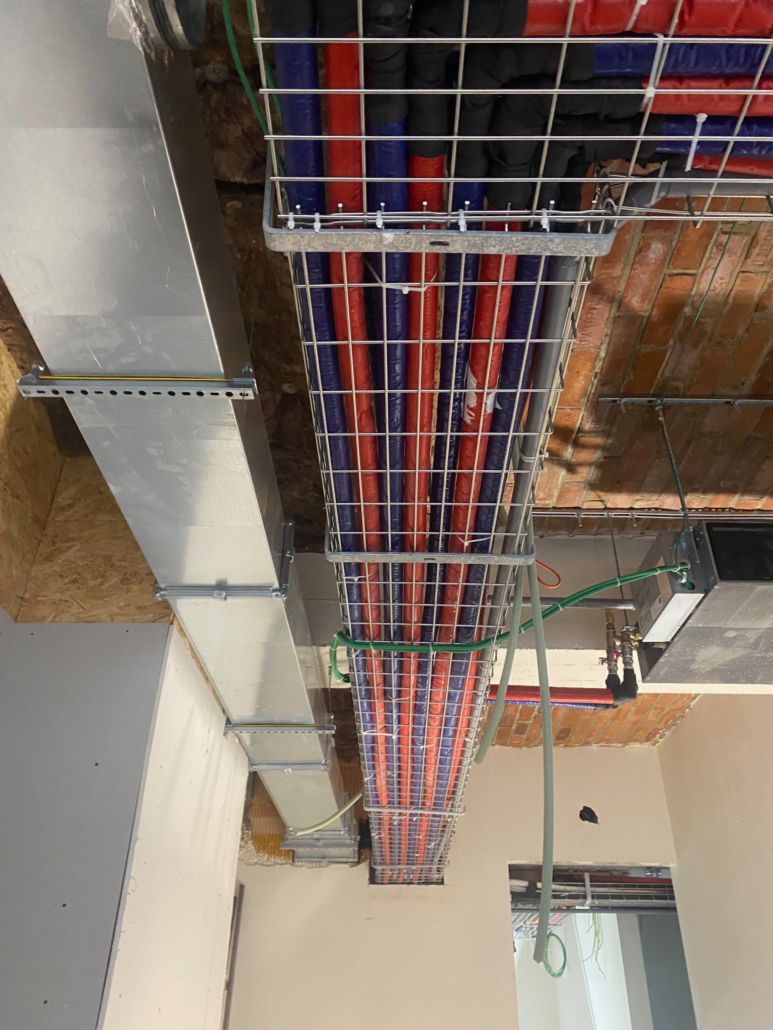 Instalacja rur grzewczych w metalowej klatce pod sufitem z cegły. Widoczny fragment wentylacji i instalacji elektrycznej. Prace instalacyjne w trakcie.