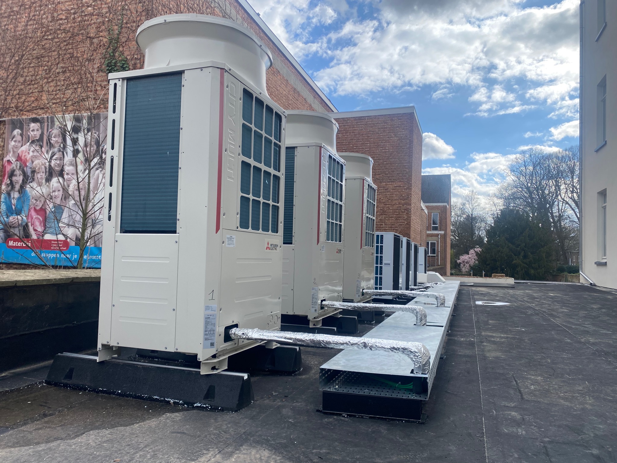 Szereg białych jednostek zewnętrznych klimatyzacji Mitsubishi Electric na dachu budynku, z widocznymi przewodami w srebrnej izolacji. Tło stanowi budynek z cegły i błękitne niebo.