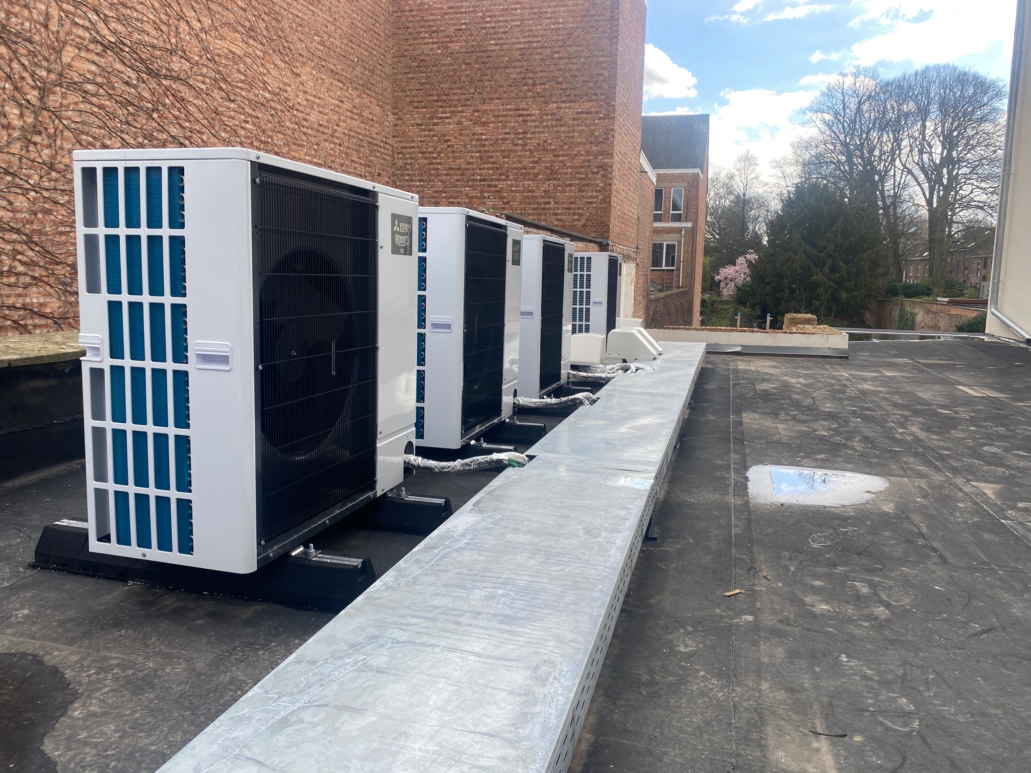 Rząd białych jednostek zewnętrznych klimatyzacji na dachu z widokiem na budynek z cegły i drzewa w tle, instalacja na czarnym dachu pokrytym papą.