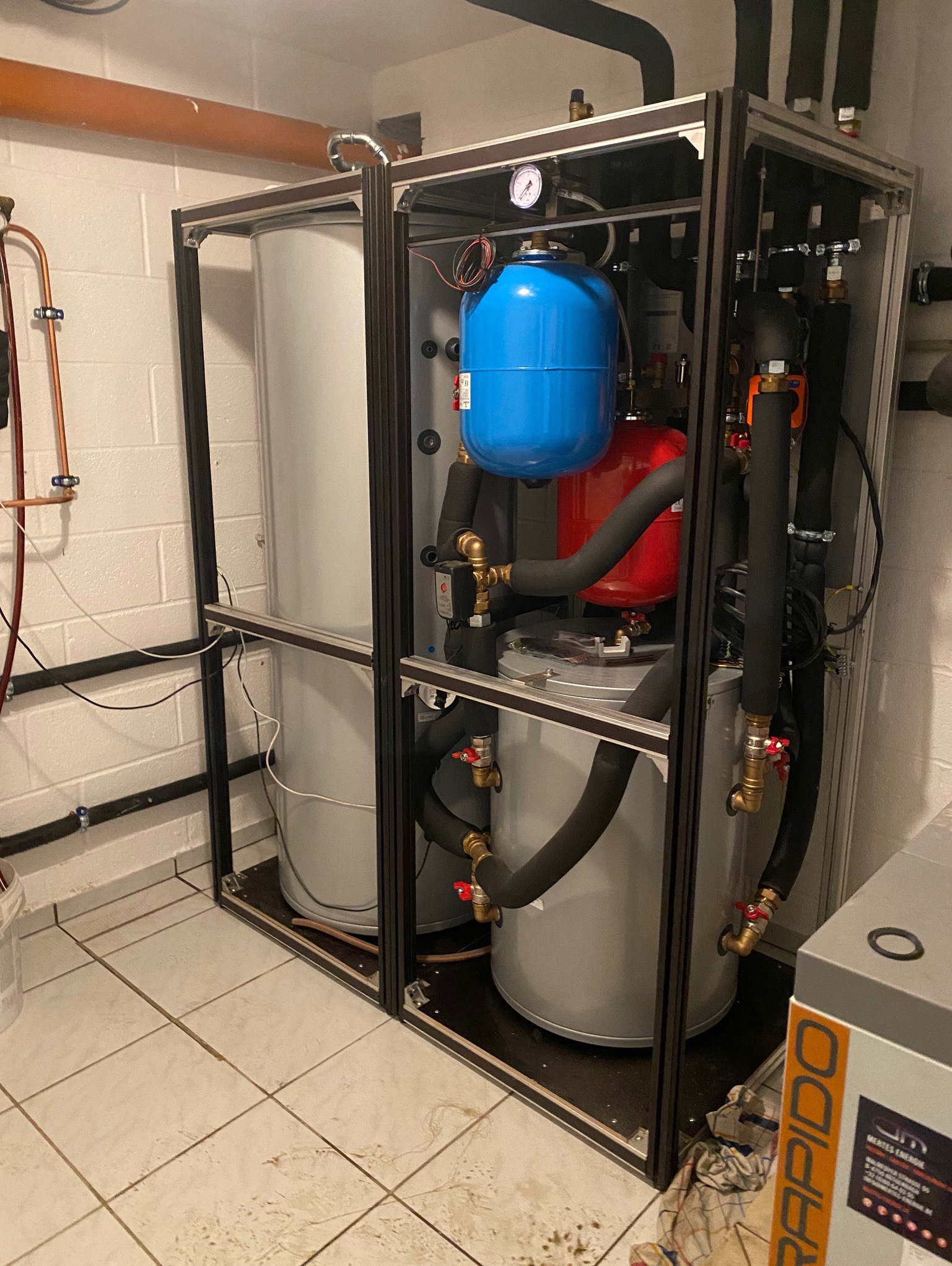 Złożona instalacja grzewcza z dwoma zbiornikami, rurami i elementami sterującymi, umieszczona w ramie z profili aluminiowych, w pomieszczeniu technicznym z białymi ścianami.