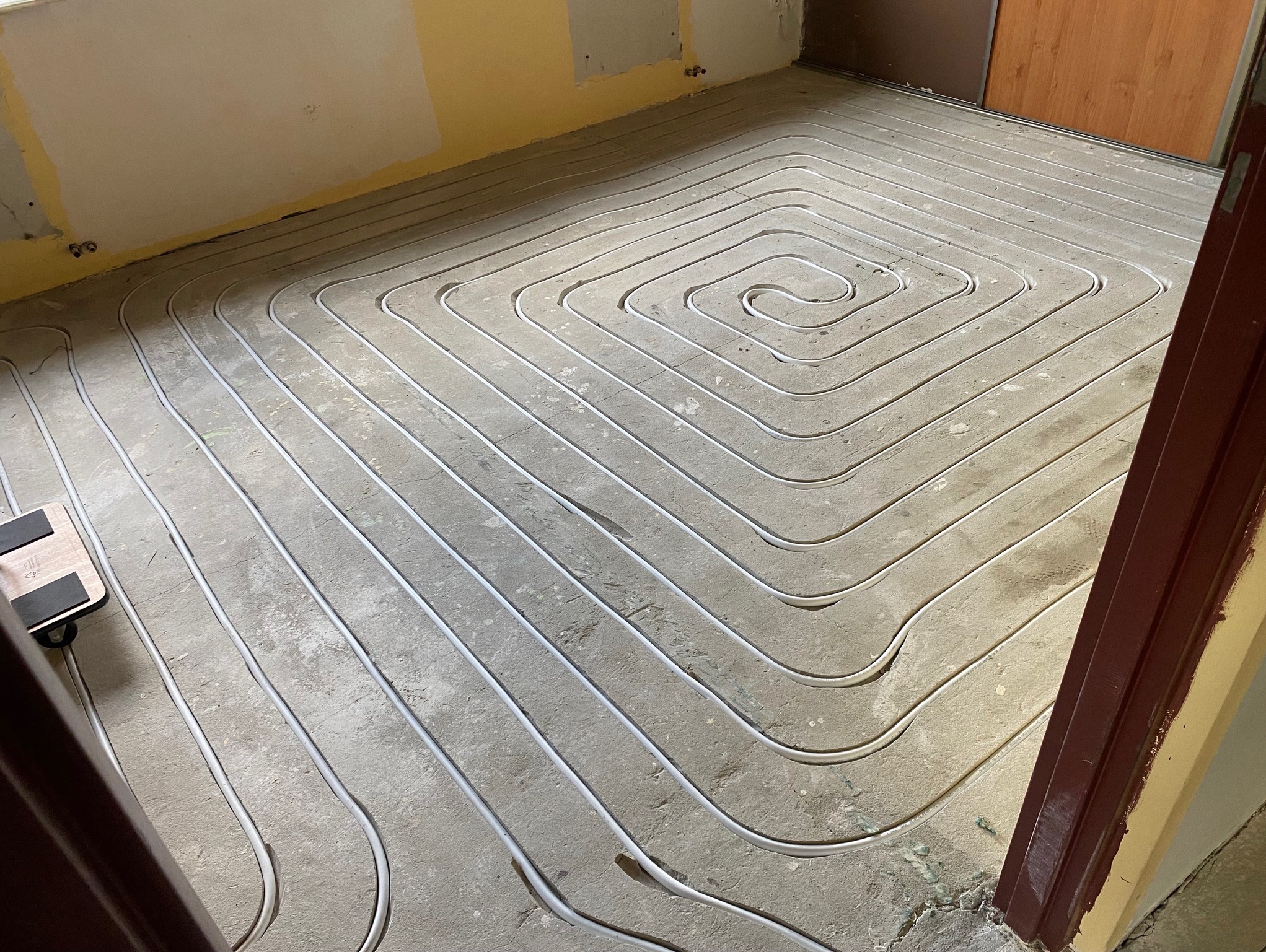 Ułożone, białe rury ogrzewania podłogowego na szarej wylewce w trakcie prac wykończeniowych. Na ścianach widoczne ślady po remoncie. Ujęcie z góry.