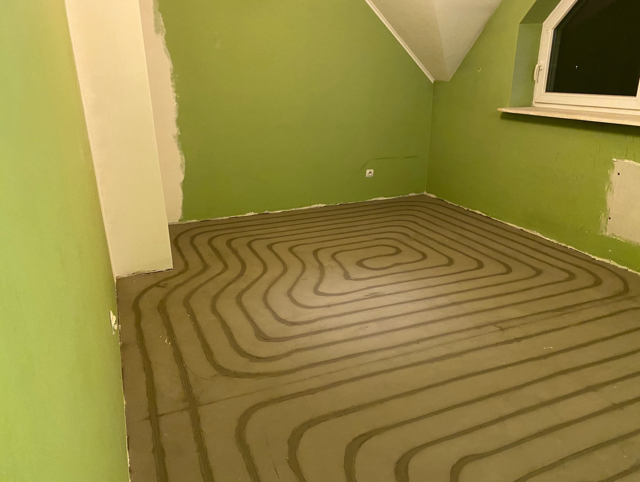 Ułożone rury ogrzewania podłogowego na wylewce w pokoju z zielonymi ścianami i oknem. Widok z góry na spiralny układ rur przed dalszymi pracami wykończeniowymi.
