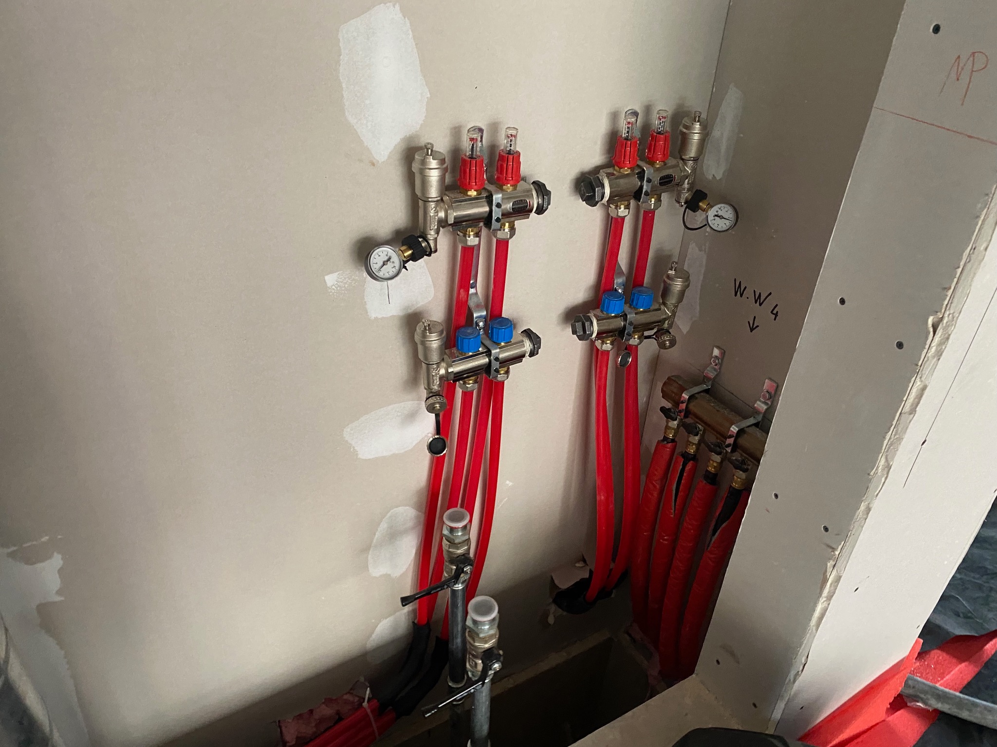 Rozdzielacz ogrzewania podłogowego z czerwonymi rurami, manometrem i zaworami, zamontowany na ścianie w surowym stanie. Widoczne napisy 'WW4' i 'MP' na ścianie.