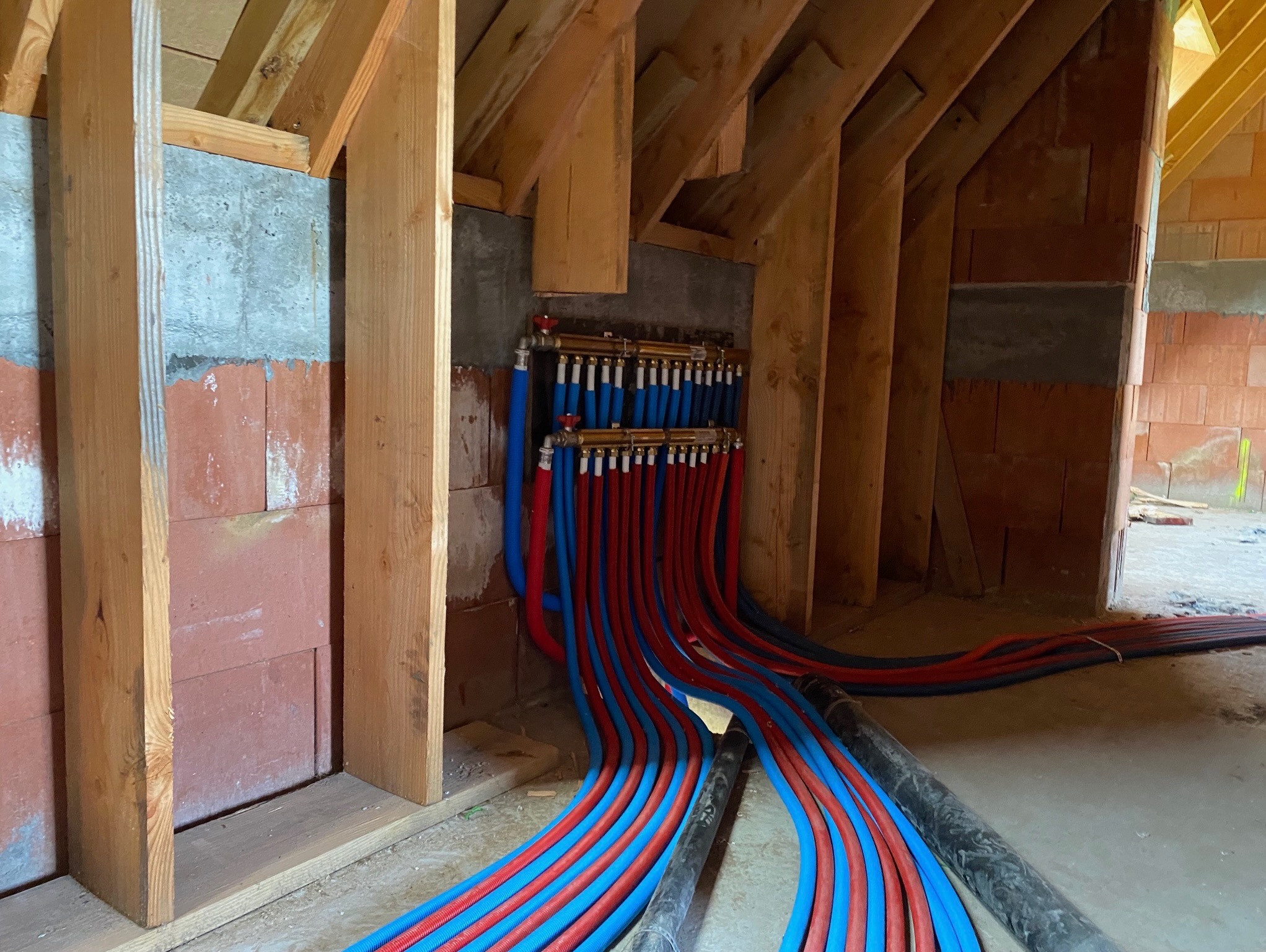 Rozdzielacz ogrzewania podłogowego z rurami PEX w kolorach niebieskim i czerwonym, zamontowany na ścianie z bloczków w surowym wnętrzu z drewnianą konstrukcją dachu.