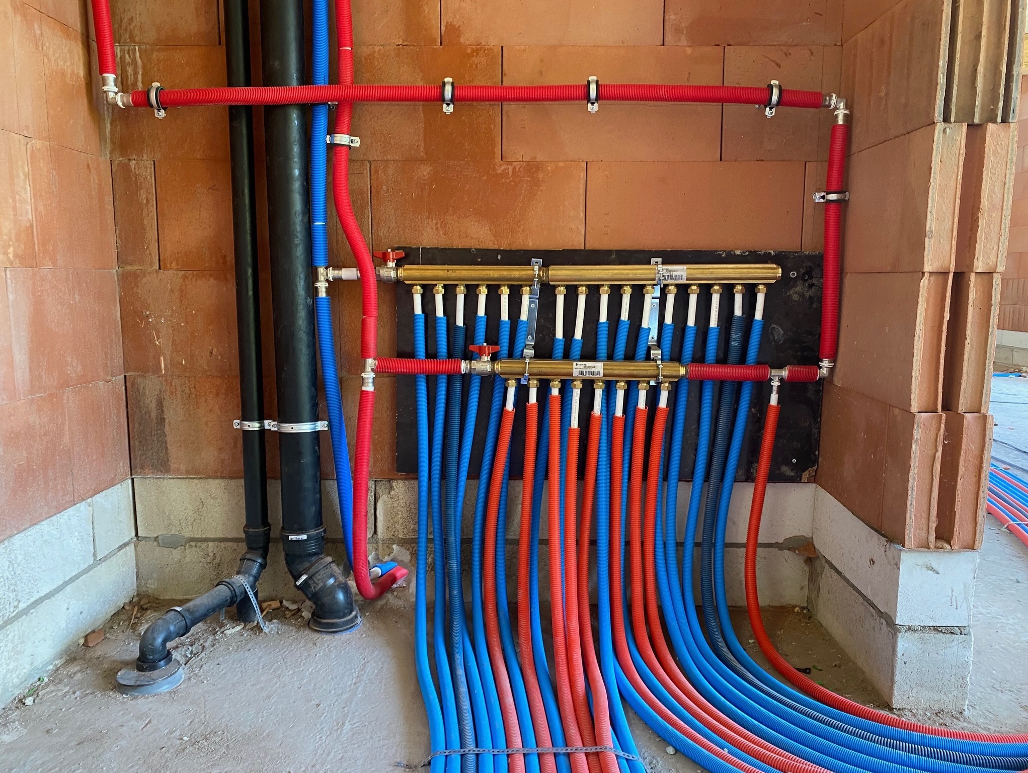 Rozdzielacz ogrzewania podłogowego z rurami w kolorach niebieskim i czerwonym, zamocowany na ścianie z cegieł. Widoczne elementy instalacji w surowym wnętrzu.