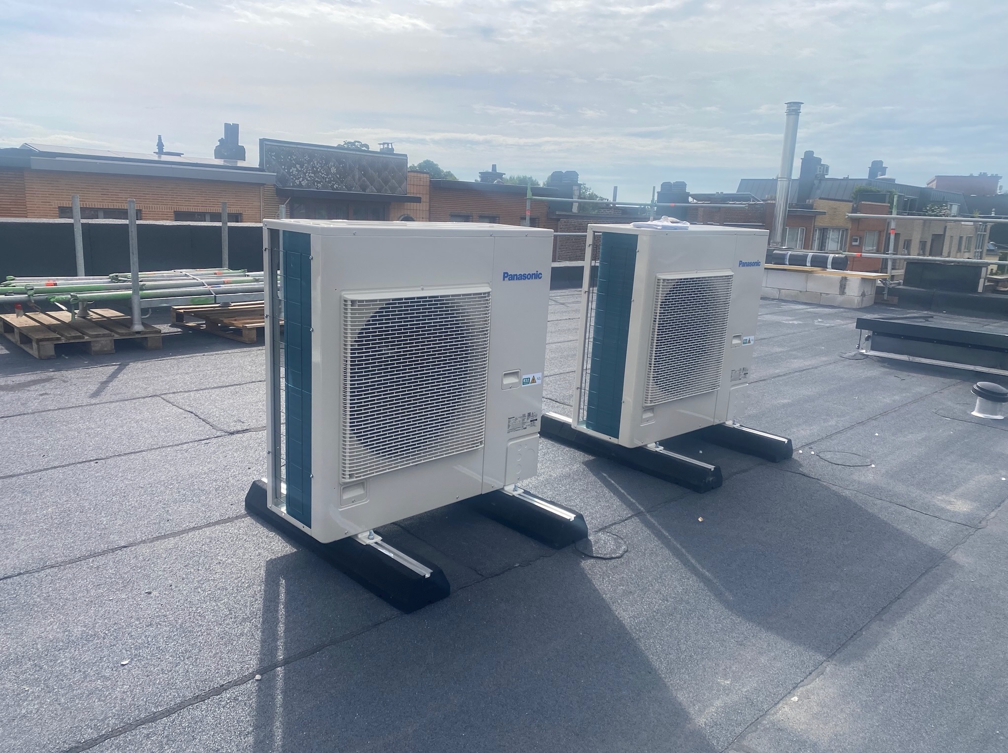 Dwa klimatyzatory Panasonic na dachu budynku, ustawione na czarnych podstawach. W tle widoczne inne budynki i elementy konstrukcyjne.