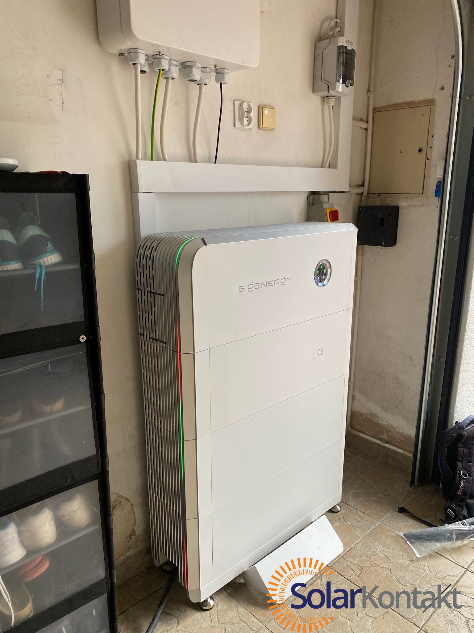Nowoczesny, biały magazyn energii SIGENergy z pionowymi wentylacjami i subtelnym zielono-czerwonym podświetleniem, podłączony do instalacji elektrycznej na ścianie w pomieszczeniu gospodarczym.