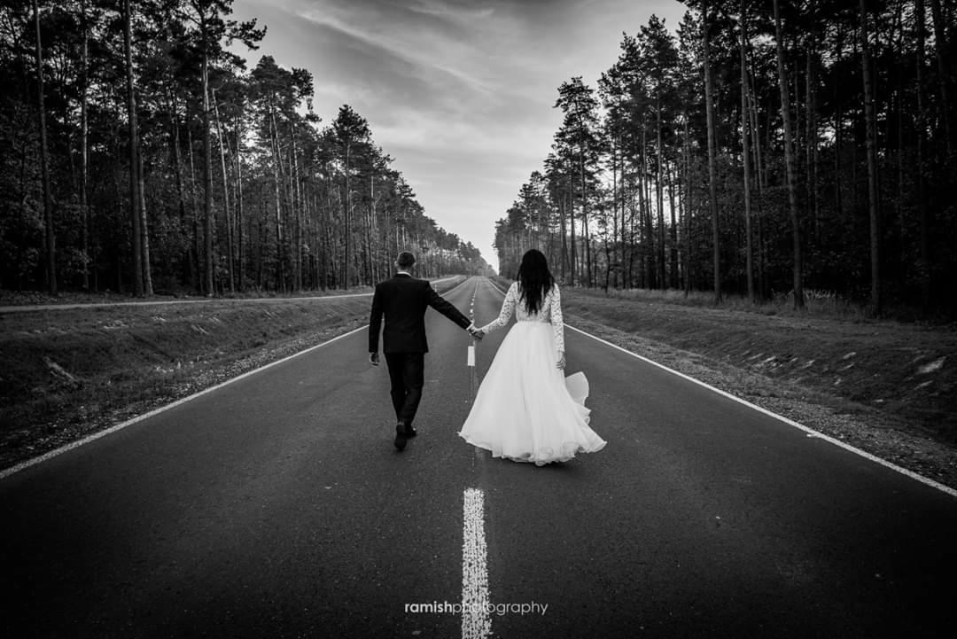 Czarno-białe zdjęcie pary młodej idącej środkiem drogi otoczonej lasem, panna młoda w sukni z długim rękawem, pan młody w garniturze, trzymają się za ręce, widok z tyłu.