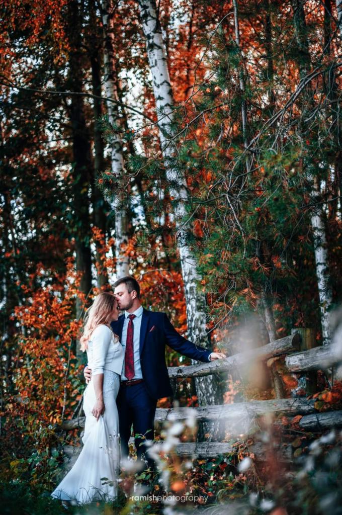 Para w eleganckich strojach pozuje w jesiennym lesie, mężczyzna całuje kobietę w czoło przy drewnianym płocie, w tle brzozy i czerwone liście.