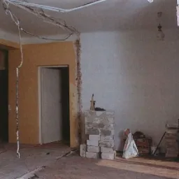 Wnętrze w trakcie rozbiórki: fragment ściany z odsłoniętą cegłą, widoczne przewody elektryczne i sterta cegieł na podłodze.