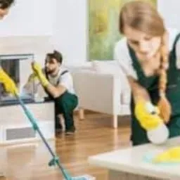 Trzy osoby w strojach roboczych sprzątają wnętrze domu: kobieta myje podłogę mopem, druga kobieta czyści powierzchnię blatu, a mężczyzna czyści obudowę kominka.