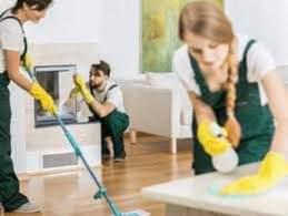 Trzy osoby w strojach roboczych sprzątają wnętrze domu: kobieta myje podłogę mopem, druga kobieta czyści powierzchnię blatu, a mężczyzna czyści obudowę kominka.