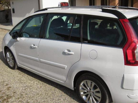 Biały samochód typu kombi z czarnym bagażnikiem dachowym i podświetlanym napisem TAXI na dachu, zaparkowany na żwirowym podjeździe przed jasnym budynkiem.