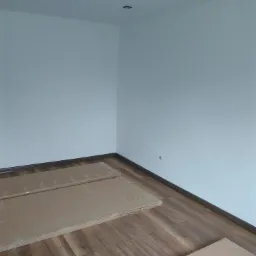 Przygotowane do montażu panele podłogowe w kartonowych opakowaniach leżą na drewnianej podłodze w pustym, pomalowanym na biało pokoju z zamontowanym oświetleniem sufitowym.